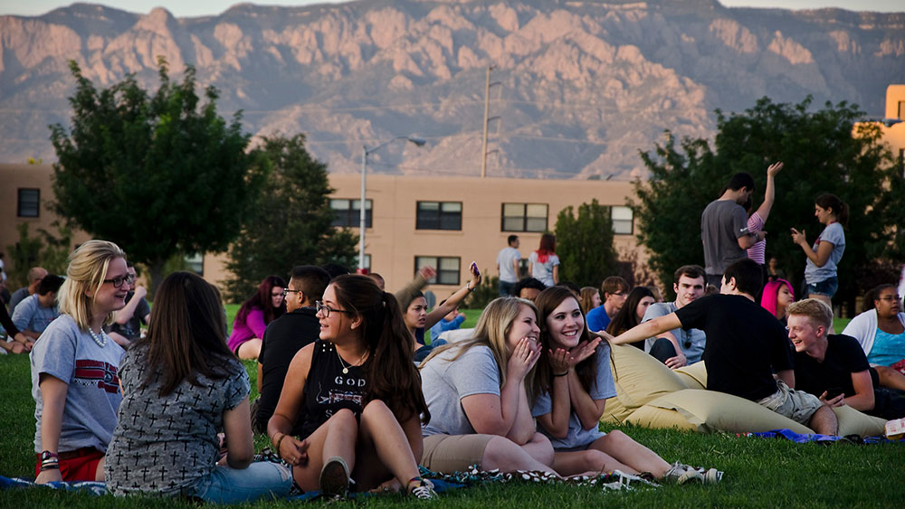 A group of students hanging out on Johnson Field with the dorms and Sandia Mountains in the background